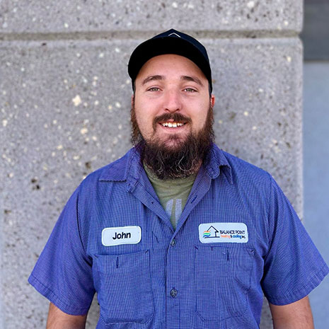 John smiling at the camera in front of a gray wall. He's wearing a blue Balance Point work shirt and a baseball cap.